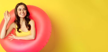 Happy young woman wearing pink swimming ring, waving hand and saying hi, enjoying vacation, summer beach holiday, standing over yellow background.