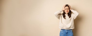 Frustrated and troubled woman facing disaster, holding hands on head in panic, looking anxious, standing over beige background.