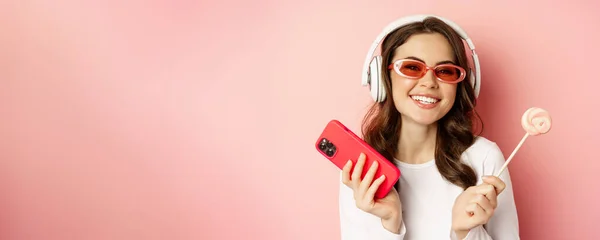 身披棒棒糖和手机 戴着太阳镜和耳机 听着音乐 站在粉色背景上的时髦年轻女子 — 图库照片