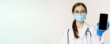 Online medical help concept. Woman doctor in glasses and face mask, showing mobile phone screen, app interface or website for patients, standing over white background.