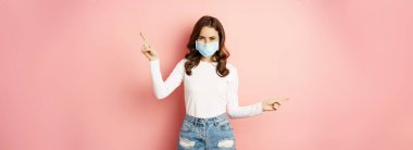 Portrait of beautiful woman in face medical mask pointing sideways, showing two choices, ways, sides, demonstrating advertisement, pink background.