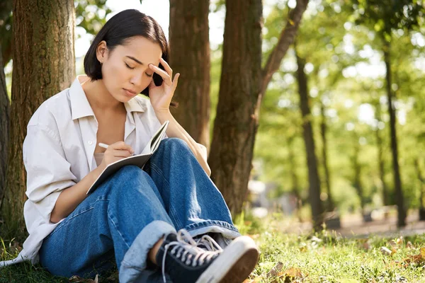 stock image Portrait of sad asian girl writing in her diary and feeling uneasy, sitting in park alone under tree, expressing her distress in notebook.