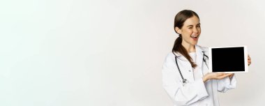 Image of woman doctor, female healthcare worker showing online medical website, digital tablet screen and smiling, standing in white coat over white background.