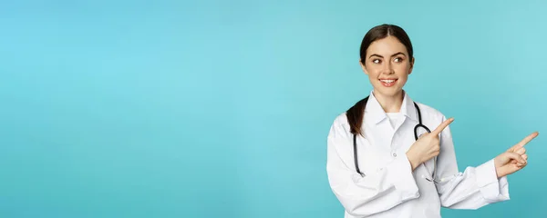 若い専門医 女性病院の労働者の肖像画 右指を指して クリニックのプロモーション契約を示し ターコイズの背景の上に立って — ストック写真