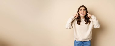 Annoyed and angry woman shut ears, complaining at loud noisy music noise, standing over beige background.
