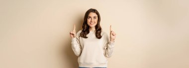 Enthusiastic girl pointing fingers up, showing announcement, promo text or logo upwards, standing over beige background.