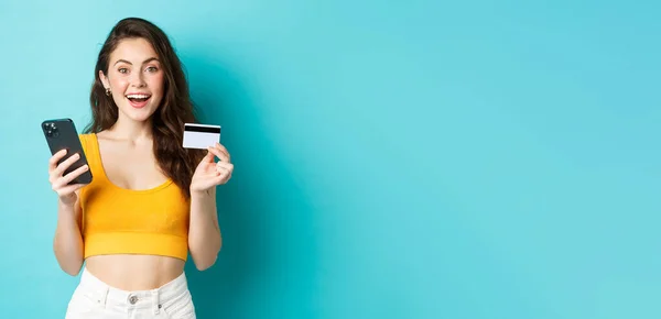 オンライン注文をしている若い幸せな女性 クレジットカードやスマートフォンでの買い物 カメラを見て驚き 青い背景の上に立って — ストック写真