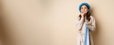 Image of modern beautiful woman posig with shopping gift bag, smiling and looking up thoughtful, thinking, standing over beige background.