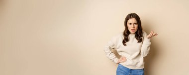 Frustrated and pissed-off woman, looking confused, arguing, standing over beige background.