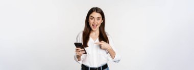 Enthusiastic girl pointing finger at smartphone and smiling, showing big sale online, using app on mobile phone, white background.