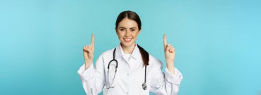 Portrait of confident young woman doctor, medical worker in coat, pointing fingers up and smiling, showing medicine advertisement, clinic promo, toquoise background.