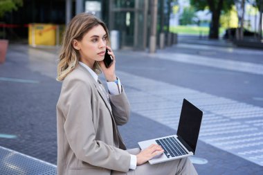 Şehir merkezinde dizüstü bilgisayarıyla oturan ve cep telefonuyla konuşan genç bir şirket kadını..