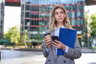 İş kadını kahve içiyor, elinde akıllı telefonuyla evrak tutuyor, ofiste çalışıyor, şehir merkezinde dışarıda duruyor..
