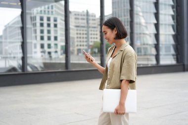 Bilgisayarlı genç Asyalı kadın, sokakta yürüyor ve mesaj atıyor, akıllı telefona bakıp gülümsüyor..