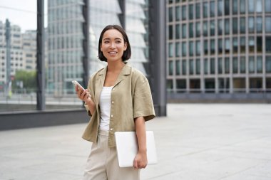 Dizüstü bilgisayarı ve akıllı telefonu olan Asyalı bir kız şehir merkezinde kameraya gülümsüyor..