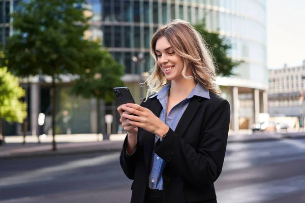 stock image Close up portrait of young corporate woman in black suit, holds smartphone, texting message while standing on street, using mobile application.