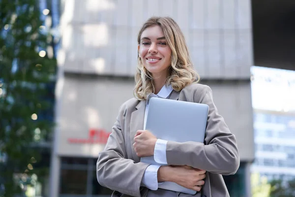 Portrait of young corporate woman, holding her laptop and smiling, going to work, wearing professional outfit, beige suite.