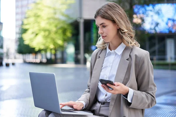 Corporate woman in suit working in city centre, using laptop and mobile phone.