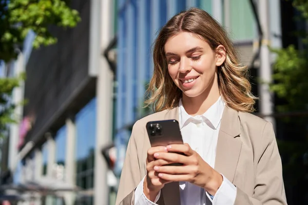 stock image Portrait of stylish businesswoman in corporate outfit, holding smartphone, standing on street and using mobile phone in hands.