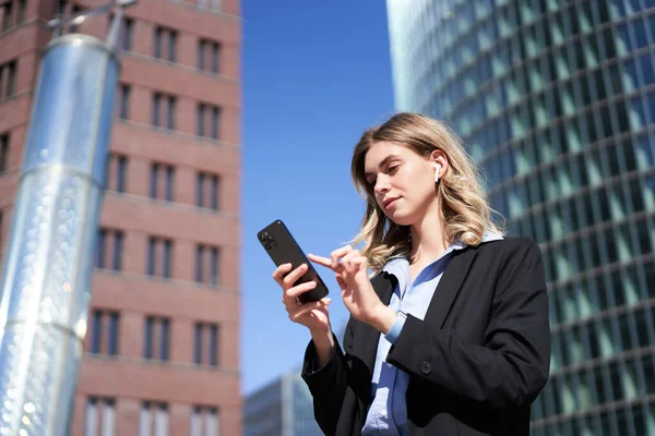 Güneşli bir günde iş kadınının elinde akıllı bir telefonla caddede dikilirken görüntüsü düşük açılı ve uygulamalı, kısa mesajlı..