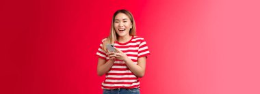 Mutlu neşeli Asyalı sarışın kız akıllı telefon kullanarak gülüyor, kız kucağında geniş bir gülümseme cihazı var, kameraya bakın eğleniyor, kız arkadaşlara mesaj atıyor, komik internet iletileri okuyor, kırmızı arka planda kaygısız duruyor..