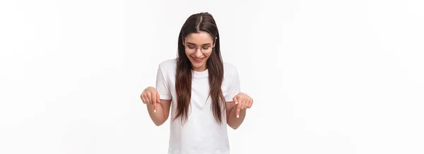 ウエストアップの肖像画の熱狂的な 笑顔幸せなブルネットの女性学生で眼鏡とTシャツ 下の広告を下を向いて見て 喜んで 白い背景を立って — ストック写真