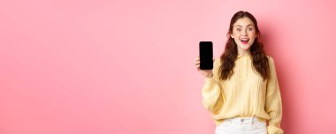 Teknoloji ve online alışveriş. Çekici kız öğrenci heyecanlı görünüyor, boş akıllı telefon ekranı gösteriyor ve gülümsüyor, pembe arka plana karşı duruyor..