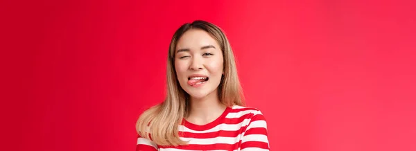 都会のアジア系女子大生の夏休みは縞模様のTシャツを着て 舌が楽しそうにカメラをウィンクして 赤い背景に立っています — ストック写真