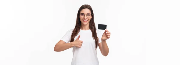 穿着T恤衫和眼镜的年轻貌美的黑发女人的形象 推荐银行 展示信用卡 满意地微笑展示大拇指 同意网上购物 建议网上购物 — 图库照片