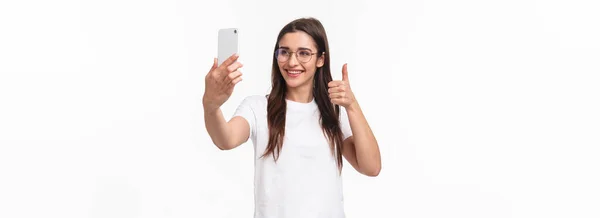 コミュニケーション テクノロジー ライフスタイルの概念 アップビート幸せの肖像画 笑顔Caucasian女の子で眼鏡 インターネットブロガー取ります自画撮り フォロワーへのビデオメッセージを記録します親指アップ 携帯電話を保持 — ストック写真