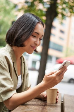 Dışarıdaki kafede oturan, kahve içen, akıllı telefon kullanan, cep telefonu okuyan Asyalı kızın portresini kapat..