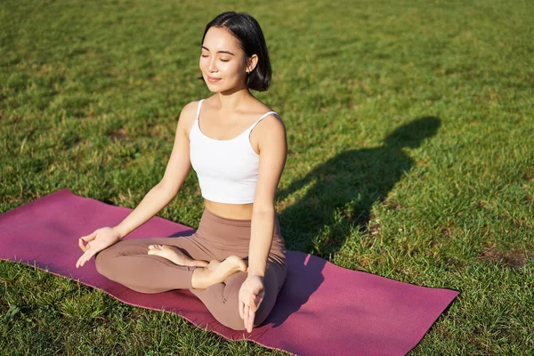 stock image Portrait of smiling asian woman meditating, doing yoga on fresh air, relaxing on rubber mat, exercising in park, breathing air, being calm.