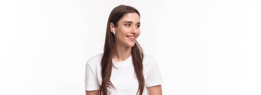 Tişörtlü, sevimli, güzel genç bir kadının yakın plan portresi, kablosuz kulaklıkla müzik dinliyor, mutlu bir gülümsemeyle başka yere bakıyor, favori podcast veya şarkı listesinin keyfini çıkarıyor, beyaz arka plan.
