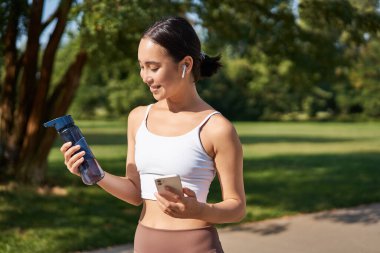 Fitness kızı portresi, kaçak içme suyu, akıllı telefona bakmak, antrenmanın ortasında parkta durmak, spor antrenmanı..