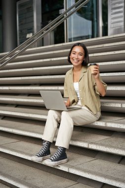 Gülümseyen kız öğrencinin dikey görüntüsü, Asyalı kadın üniversite kampüsünün merdivenlerinde oturuyor ve kahve içiyor, ödevlerini dizüstü bilgisayarında yapıyor..
