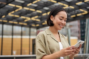 Teknoloji insanları. Genç gülümseyen Asyalı kız akıllı telefon tutuyor, sokaklarda dolaşırken cep telefonu uygulaması kullanıyor..