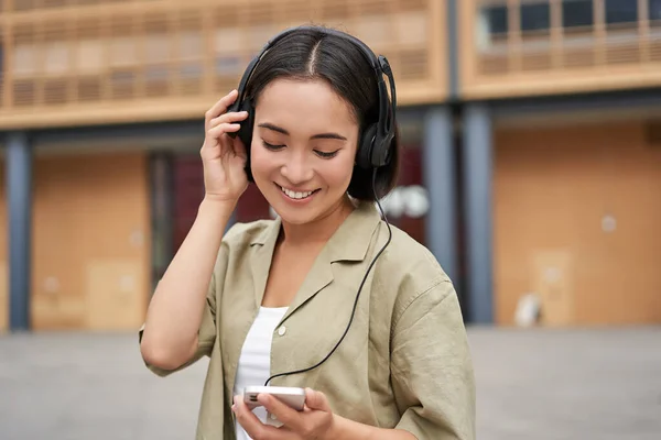 Streaming services concept. Happy asian girl listens music in headphones, holds mobile phone, choosing track or podcast, walking on street.