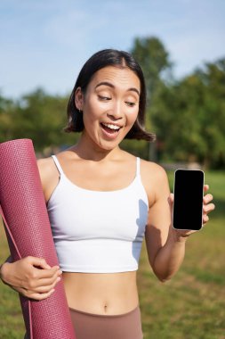 Heyecanlı fitness kızı spor ve egzersiz için başvuru öneriyor, telefon ekranını gösteriyor, antrenmandan sonra parkta kauçuk yoga minderiyle duruyor..