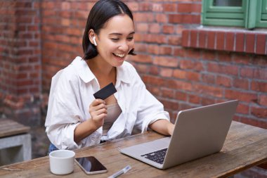 Bilgisayarlı ve kablosuz kulaklıklı gülümseyen Asyalı kadın, kredi kartıyla ödeme yapıyor, internetten alıyor, bir fincan kahveyle oturuyor..