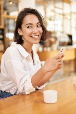 Cafede oturan, kahve içen ve akıllı telefon kullanan Asyalı bir kadının dikey portresi. Yaşam tarzı ve insan kavramı