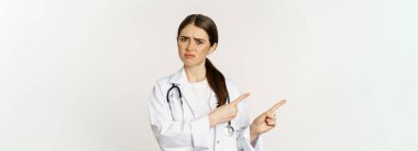 Sad and disappointed female doctor, frowning frustrated, pointing fingers and complaining, standing in white coat over white background.