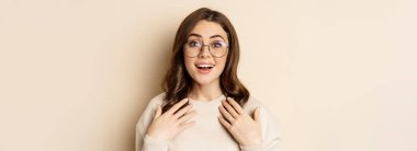 Close up portrait of beautiful girl in glasses, looking amazed and surprised, gasping excited, standing over beige background.