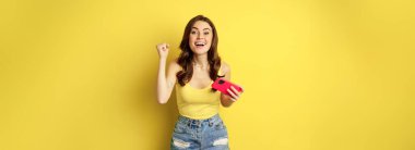 Enthusiastic young beautiful girl holding smartphone horizontally, rejoicing, winning on mobile phone video game, standing against yellow background.
