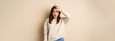 Woman showing loser sign on forehead, L word, standing over beige background. Copy space