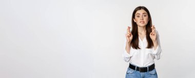 Hopeful woman praying, cross fingers for good luck, begging, yearning smth, standing over white background. Copy space