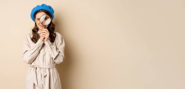 Concept of search. Young stylish girl in detective clothing, looking through magnifying glass, big eye, searching, looking for something, standing over beige background.