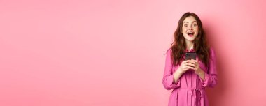 Şaşkın bakışlı, nefes nefese kalmış güzel esmer kız elinde akıllı bir telefonla harika haberleri internetten izliyor, pembe arka planda duruyor..