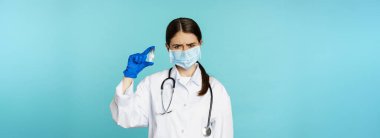 Üzgün ve hayal kırıklığına uğramış maskeli kadın doktor, aşıyı üzgün bir suratla gösteriyor, mavi arka planda üniformalı..