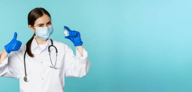 Gülümseyen doktor portresi, tıbbi personel yüz maskesi ve lastik eldiven, baş parmak ve koronavirüs gösteren, omikron aşısı, mavi arka planda duran.