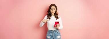 Cute girl pointing finger at smartphone with curious face expression, have you seen this gesture, showing online shopping offer on mobile phone app, pink background.